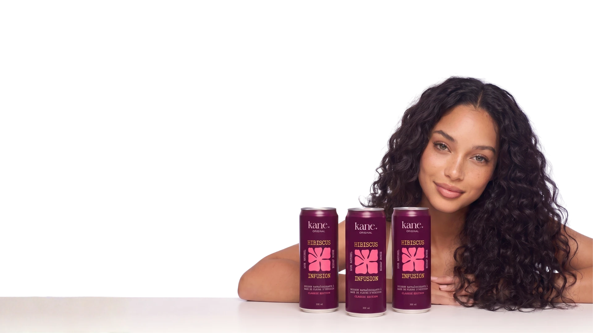 Une femme souriante aux cheveux bouclés rapproche trois canettes Kane Original Hibiscus Infusion